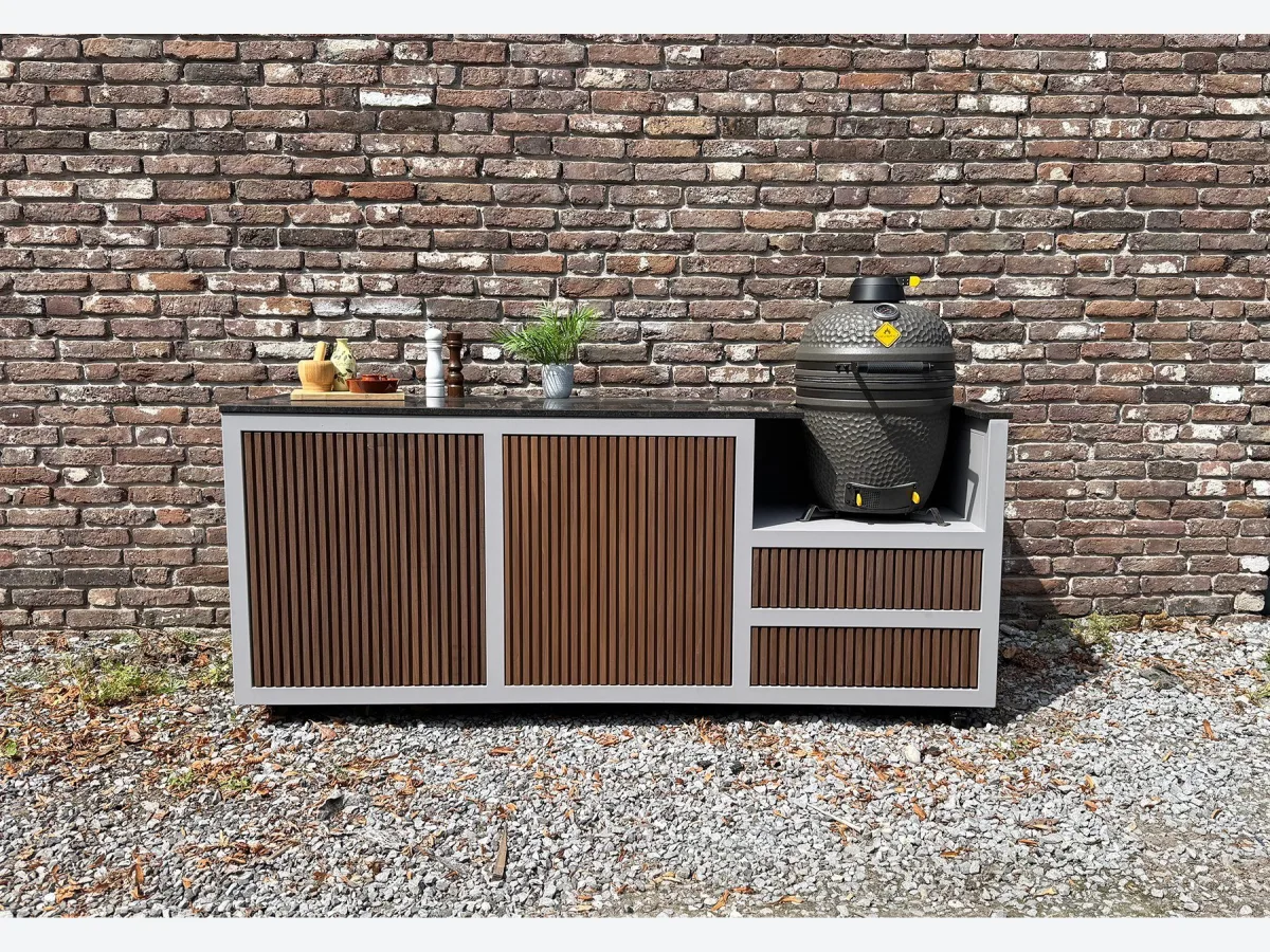 HoutenmeubelsXL Bandung, Buitenkeuken Bandung van thermohout met grijs stalen frame en natuurstenen blad, met kamado rechts, recht van voren voor een bakstenen muur