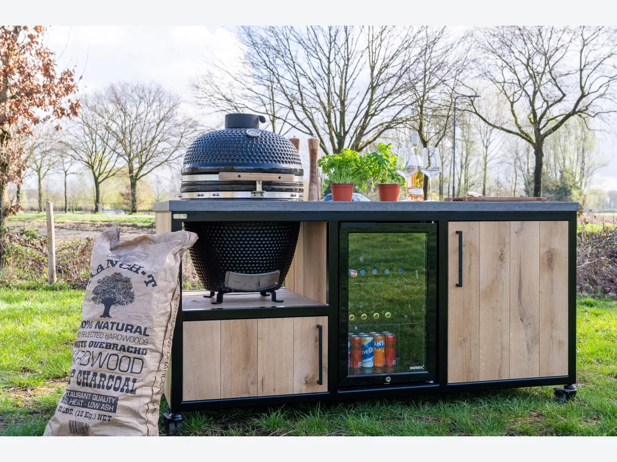 MB Industrial Living Basic, Buitenkeuken Basic op wielen met kamado links, inbouwkoelkast en houten deur, in het gras met bomen op de achtergrond