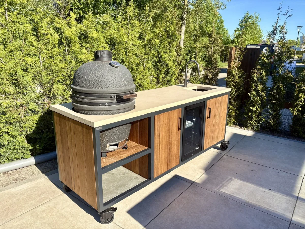 MB Industrial Living Maatwerk buitenkeuken, Maatwerk buitenkeuken op wielen met kamado links, spoelbak en inbouwkoelkast, driekwart van linksvoor op een terras met coniferen
