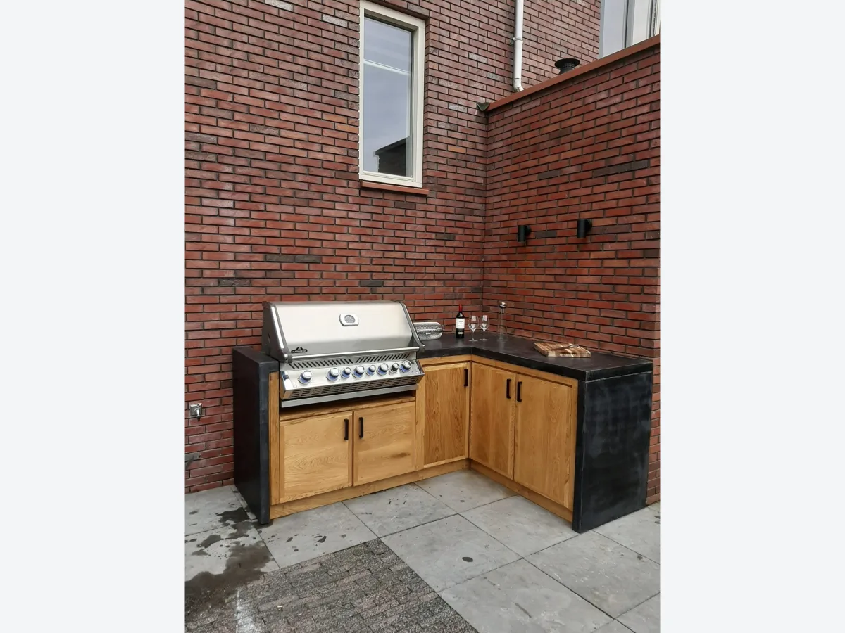 Pronck Buitenkeuken Beton, Betonnen buitenkeuken in L-vorm met eiken kastdeuren en ingebouwde Napoleon gasbarbecue tegen een bakstenen gevel