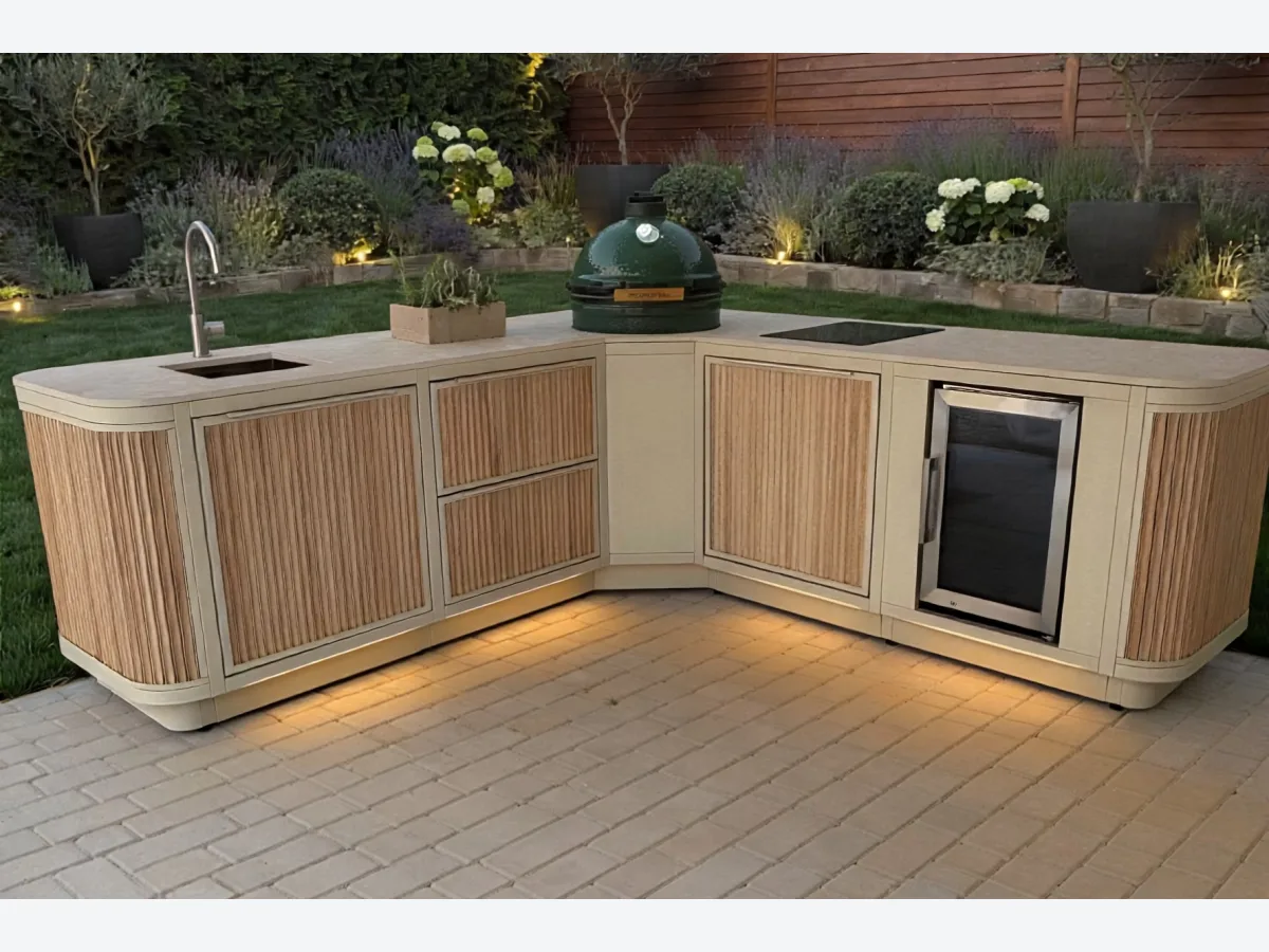 Rêveluxe Sur Mesure, Render van een Sur Mesure hoekopstelling in beige met eiken rondingen, Big Green Egg, spoelbak, kookplaat en koelkast in een tuin met plintverlichting