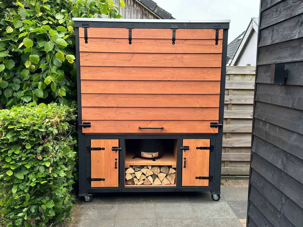 Stilt Close, Buitenkeuken Close met gesloten klep en open haardhoutvak, in een tuin tegen een zwarte wand