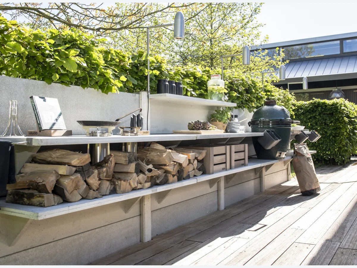 WWOO Beton, WWOO betonnen buitenkeuken in grijs langs een beukenhaag op een houten terras, met Big Green Egg, haardhout en lampen