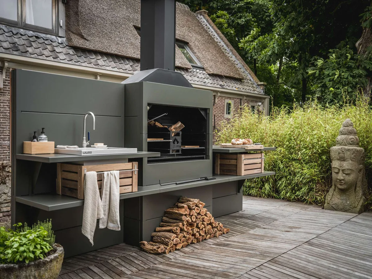 WWOO Staal, WWOO stalen buitenkeuken in donkergroen met ingebouwde braai, spoelbak en haardhout, op een houten vlonder bij een rietgedekt huis